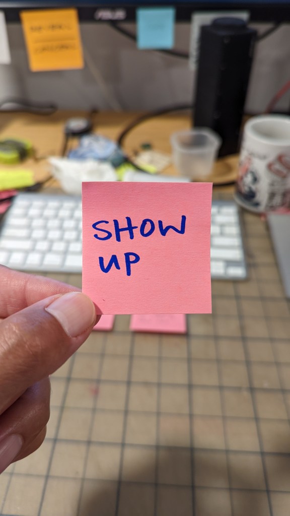 "Show up" written with a blue sharpie on a pink post it. Held between thumb and fore finger with my messy desk in the background.